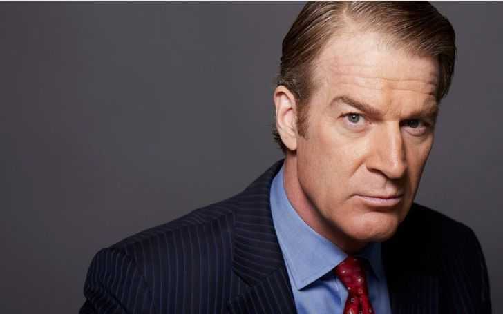Kevin Kilner in a black suit in a serious face looks at the camera.
