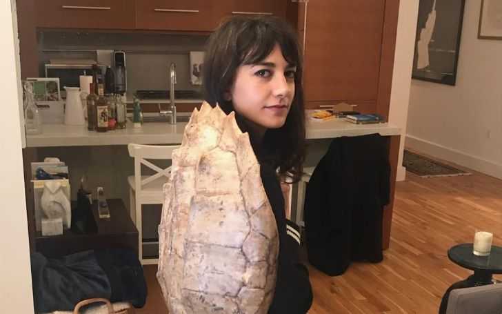 Sheila Vand looks at the camera in a turtle shell on her back.